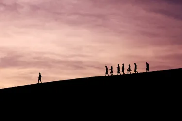 The silhouette of 8 people descending a hill at sunset with a purple sky. One person is out in front, ahead of the rest of the group.