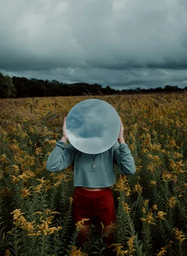 A person in a field of flowers holding a sphere up to their head that is reflecting the cloudy sky above