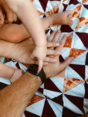 Two adult hands with a baby's hand and and leg sitting on top. They are all on a quilt with brown and white triangle shapes on it.