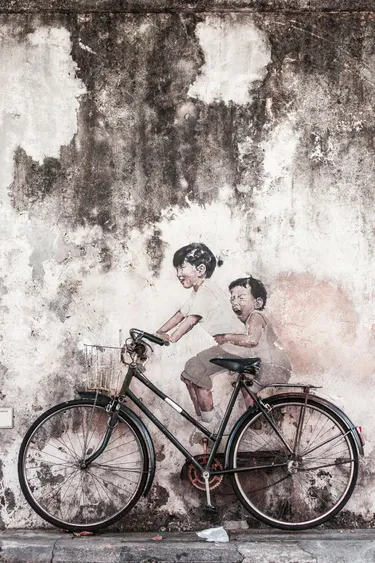 A bicycle leaning against an old, stained wall with a painting of two children on the wall. The bicylce is place in such a way that it looks like the children are riding the bike.