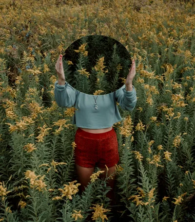 A woman standing in a field of yellow flowers wearing a teal jumper and red shorts holding a mirror up in front of her face which is reflecting the yellow flowers.
